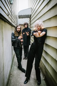 three people in black outfits holding sparklers on the sidewalk next to a wall and building