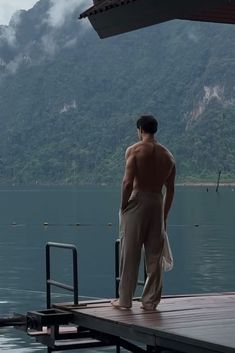 a shirtless man standing on a dock looking out at the water with mountains in the background