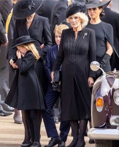 two women in black dresses and hats are standing next to a car with other people behind them