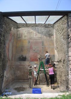 three people working on an ancient painting in a stone room with green scaffolding