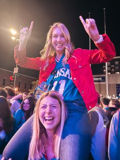 two women sitting on the shoulders of each other at a sporting event with their hands in the air