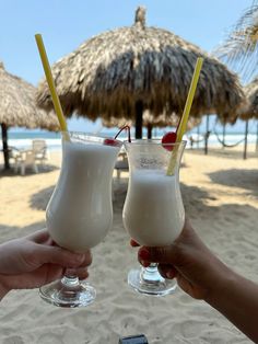 two people holding up glasses with drinks on the beach in front of straw umbrellas