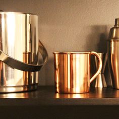 two shiny metal mugs sitting on top of a wooden shelf next to a shaker