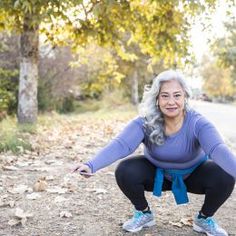 an older woman squatting down on the ground in front of some trees and leaves