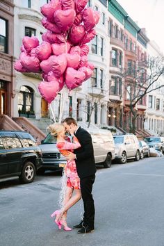 a man and woman kissing in the street with pink balloons attached to their heads as they stand next to each other