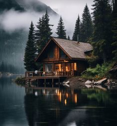 a cabin sits on the shore of a lake surrounded by pine trees and foggy mountains