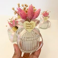 a hand holding a small glass bottle with pink flowers in it and a heart shaped label on the top