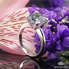 a ring with a princess cut diamond on it and purple flowers in the foreground