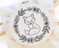 a hand embroidered fox sitting in a floral frame on a white linen background with a wooden hoop