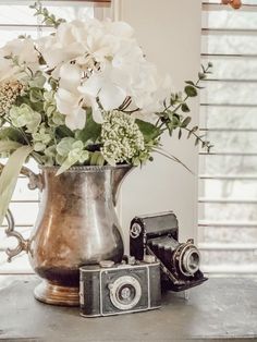 a silver pitcher with white flowers in it and an old camera sitting on a table