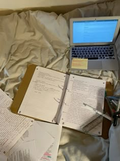 an open laptop computer sitting on top of a bed next to sheets of paper and pens