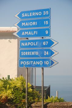 blue street signs pointing in different directions on the side of a building near the ocean