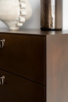 a brown dresser with two drawers and a white vase on the wall in the background