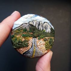 a hand holding a painted rock with trees and mountains in the background, on top of a black surface