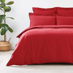 a bed with red sheets and pillows in a room next to a potted plant