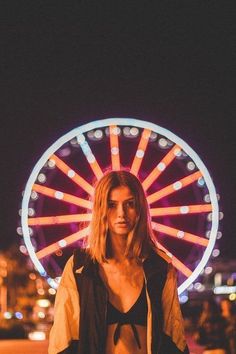 a woman standing in front of a ferris wheel at night with the lights on behind her