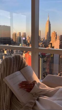 an open book sitting on top of a bed in front of a window overlooking a city