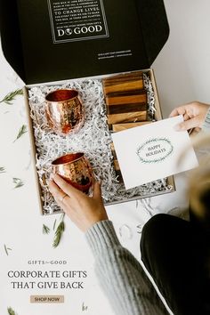 a person holding two copper mugs in a box with the words corporate gifts that give back written on it