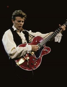 a man with a red guitar in his right hand and black vest on, playing the guitar