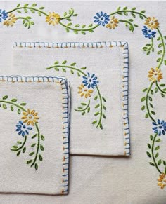 three embroidered placemats with flowers and leaves on white linen, one in blue and the other in yellow