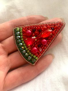 a hand holding a red and green brooch with beads on it's side