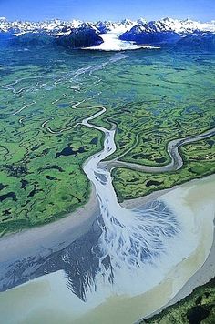 an aerial view of a river running through the middle of a lush green field with snow capped mountains in the distance