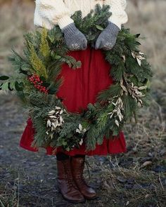 a woman in a red skirt and white sweater holding a wreath with evergreens on it