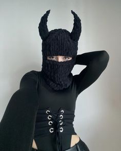 a woman wearing a knit devil mask and black outfit with horns on her head, standing in front of a white wall