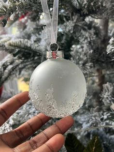 a hand holding an ornament in front of a christmas tree with snow on it