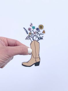 a hand holding up a sticker with flowers in a cowboy boot on it's side