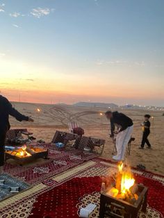 two men standing around a campfire on the beach at sunset with people in the background