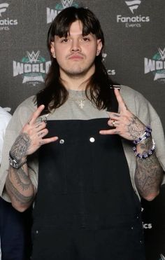 a man with long hair and tattoos standing in front of a wall holding his hands up