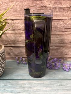 the tumbler cup has purple flowers in it and is next to a potted plant