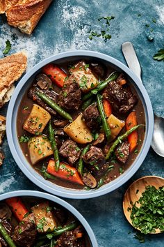 two bowls filled with beef and vegetables next to bread on a blue counter top,