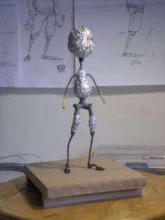 a metal figure standing on top of a wooden table in front of two drawings behind it