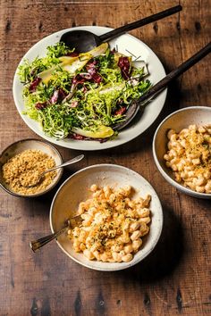 three bowls filled with macaroni and cheese next to a salad on a wooden table