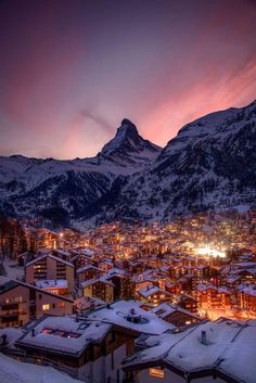 snow covered mountains and houses at night in the foreground are lit up by bright lights