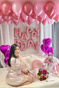 a woman sitting on top of a bed next to pink balloons and presents in front of her