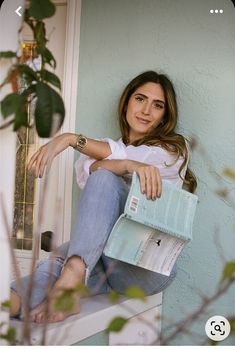 a woman sitting on a window sill holding a blue purse and looking at the camera