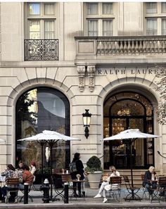 people sitting at tables in front of a building with white umbrellas on the sidewalk