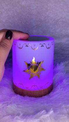 a hand holding a lit candle in front of a purple glass cup with an ornate design on it