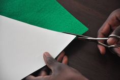 someone cutting paper with scissors on top of a wooden table next to green and white papers