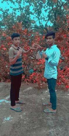 two young men standing next to each other in front of some bushes and red flowers