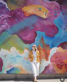 a woman standing in front of a colorful wall with flowers painted on it and wearing white pants