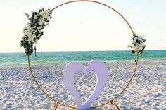 a heart shaped sign on the beach with flowers and greenery in front of it