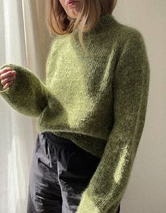 a woman standing in front of a window wearing a green sweater and black pants with her hand on her hip