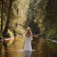 a woman in a white dress is standing in the middle of a river surrounded by trees