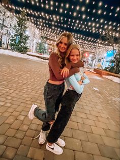 two young women hugging each other on a brick walkway with christmas lights in the background