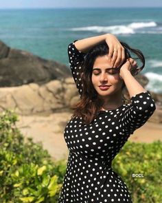 a woman in a black and white polka dot dress stands near the ocean with her hands on her head