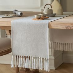 a white table runner with tassels and cups on it in front of a dining room table